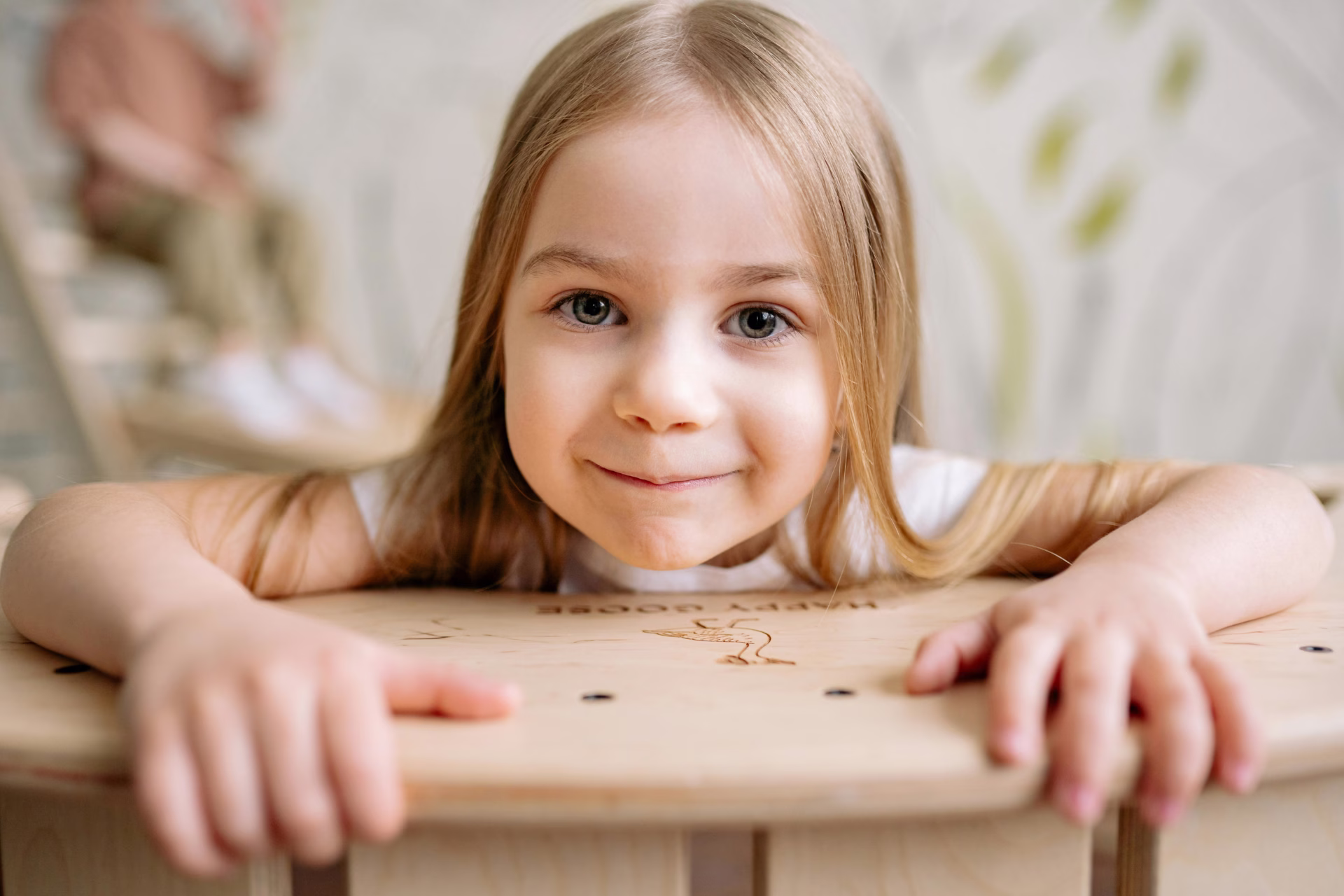 An autistic twin girl with blonde hair smiles at the camera while leaning on a table.
