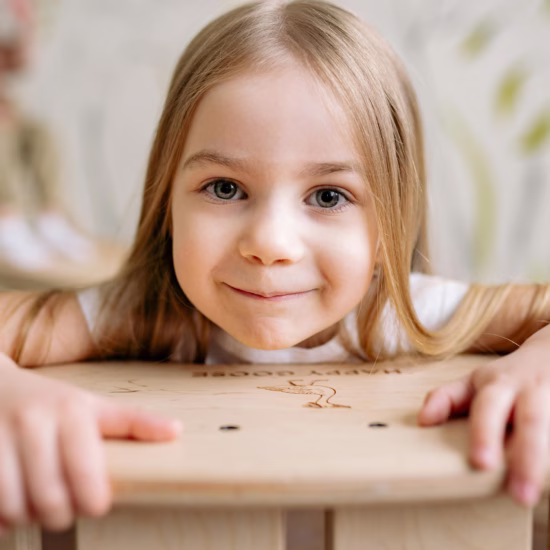 An autistic twin girl with blonde hair smiles at the camera while leaning on a table.