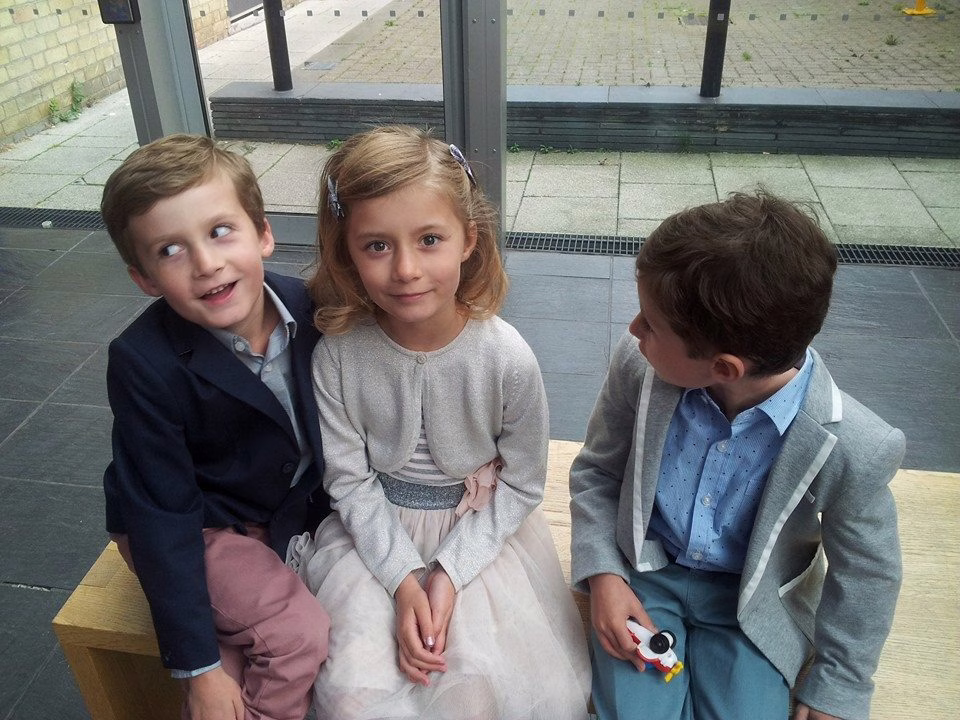 Three children (triplets) sit on a bench outside a wedding venue. Only one child is looking at the camera. 
